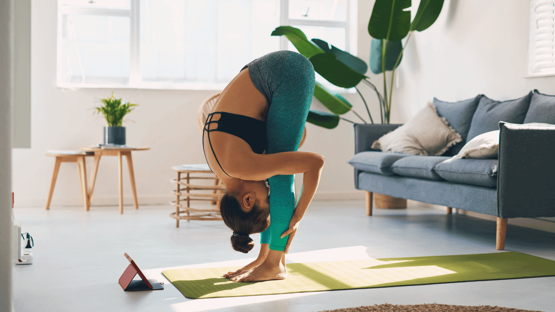Woman in athletic wear folding forward into a hip hinge on a green mat at home, holding her legs tight against her torso.