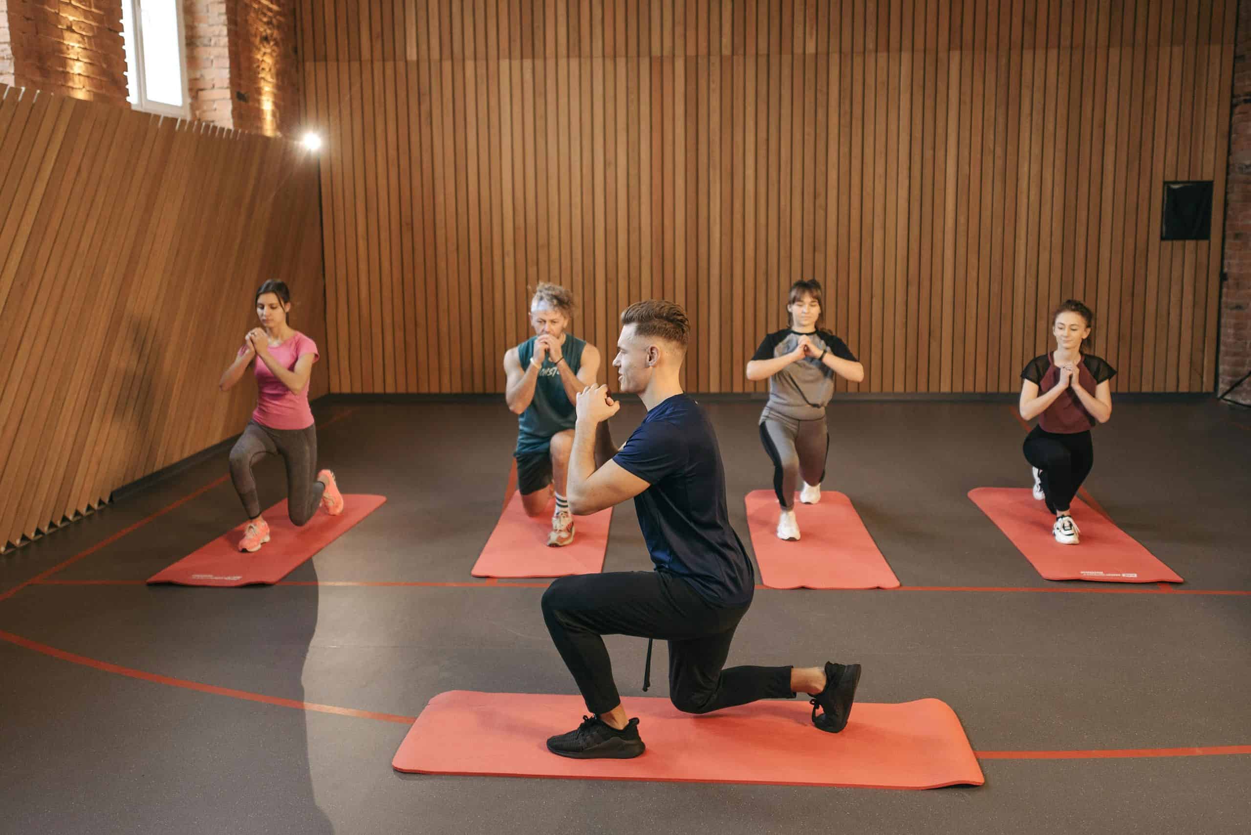 pexels-pavel-danilyuk-6339347 A fitness instructor leads a class of four on red mats, all lunging in sync with hands clasped at chest in a wood-paneled studio.