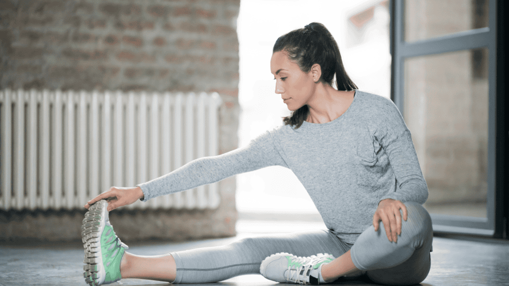 Active lady wearing sweatshirt and pants doing seated hamstring stretch at home