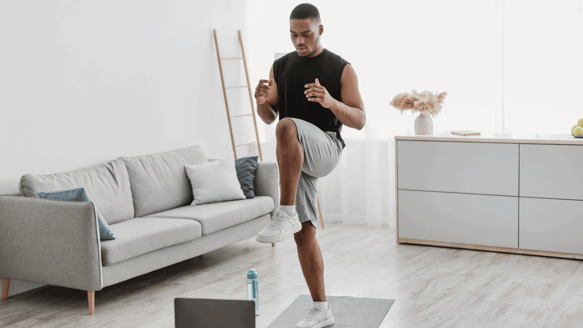Man in athletic wear performing standing knee drives on a yoga mat at home, lifting one knee up towards his chest, with a water bottle and laptop nearby.
