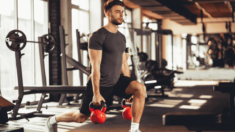 Man performing a lunge exercise with red kettlebells in a gym, showcasing a beginner-friendly HIIT workout for men.