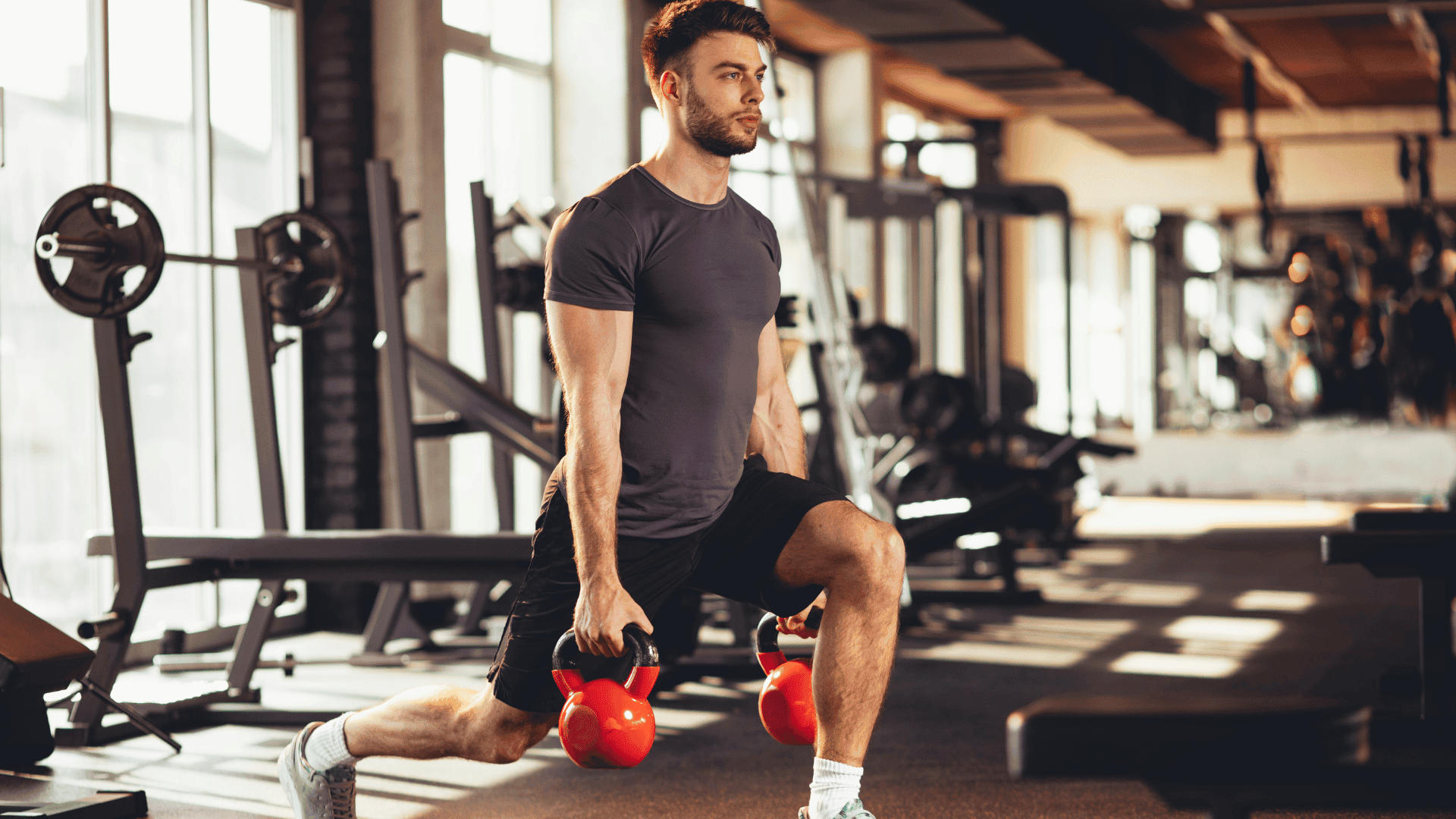 Man performing a lunge exercise with red kettlebells in a gym, showcasing a beginner-friendly HIIT workout for men.