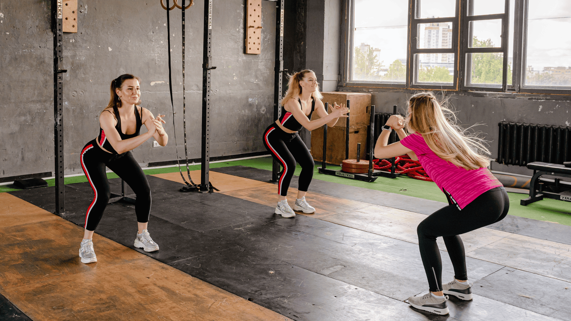 Three women performing bodyweight squats together in a gym, demonstrating a beginner-friendly lower body exercise with no equipment.