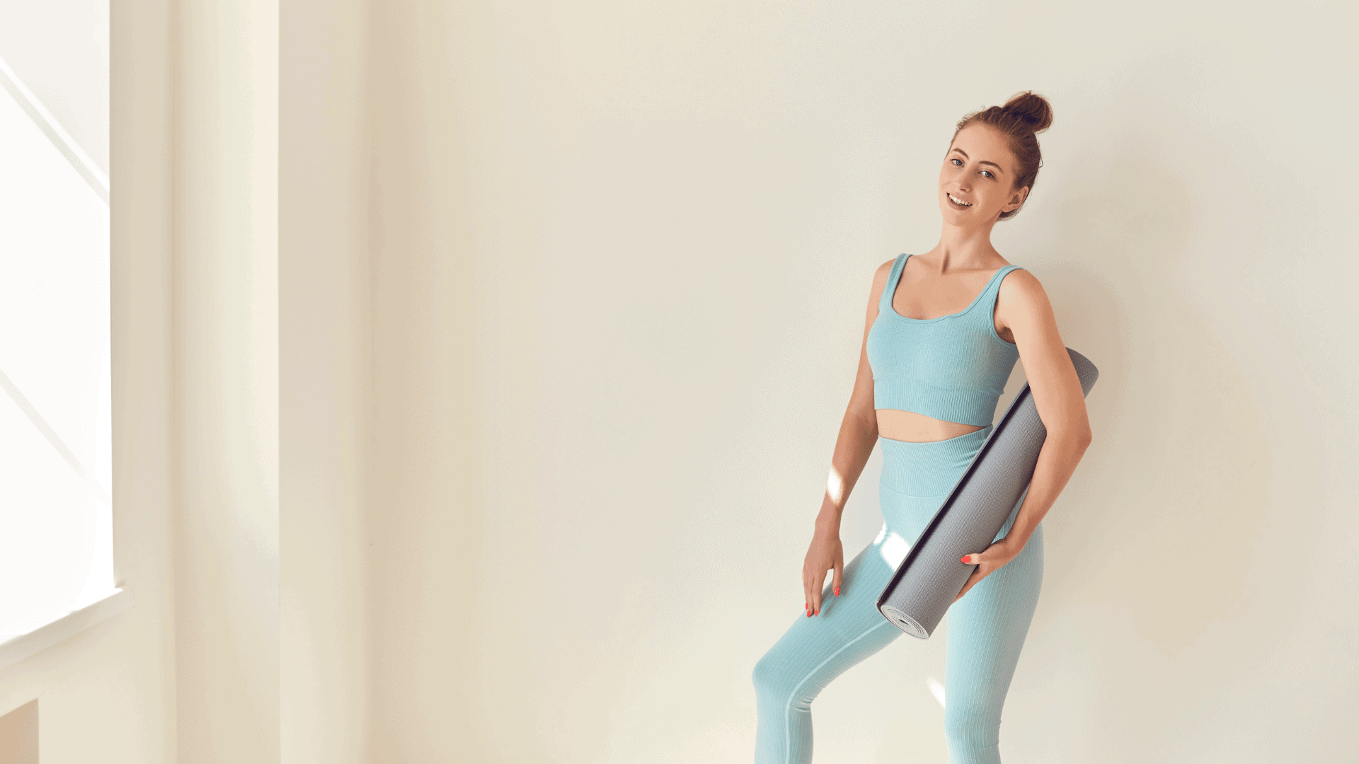 Woman in blue workout clothes holding a yoga mat, standing in a bright, minimal home Pilates space.