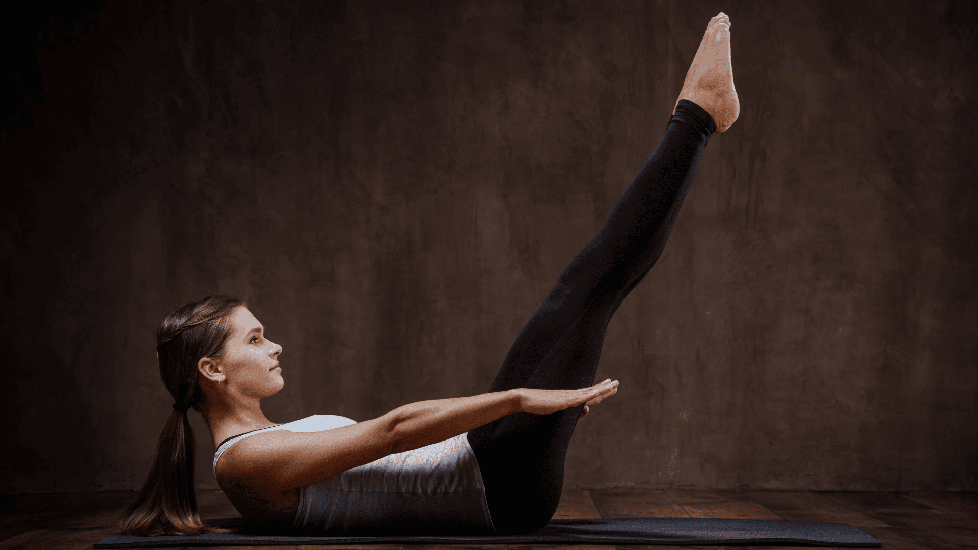Woman performing The Hundred Pilates exercise on a mat against a brown background