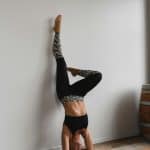 Woman in black and patterned leggings and black sports bra performing a headstand against a white wall on wooden floor indoors.