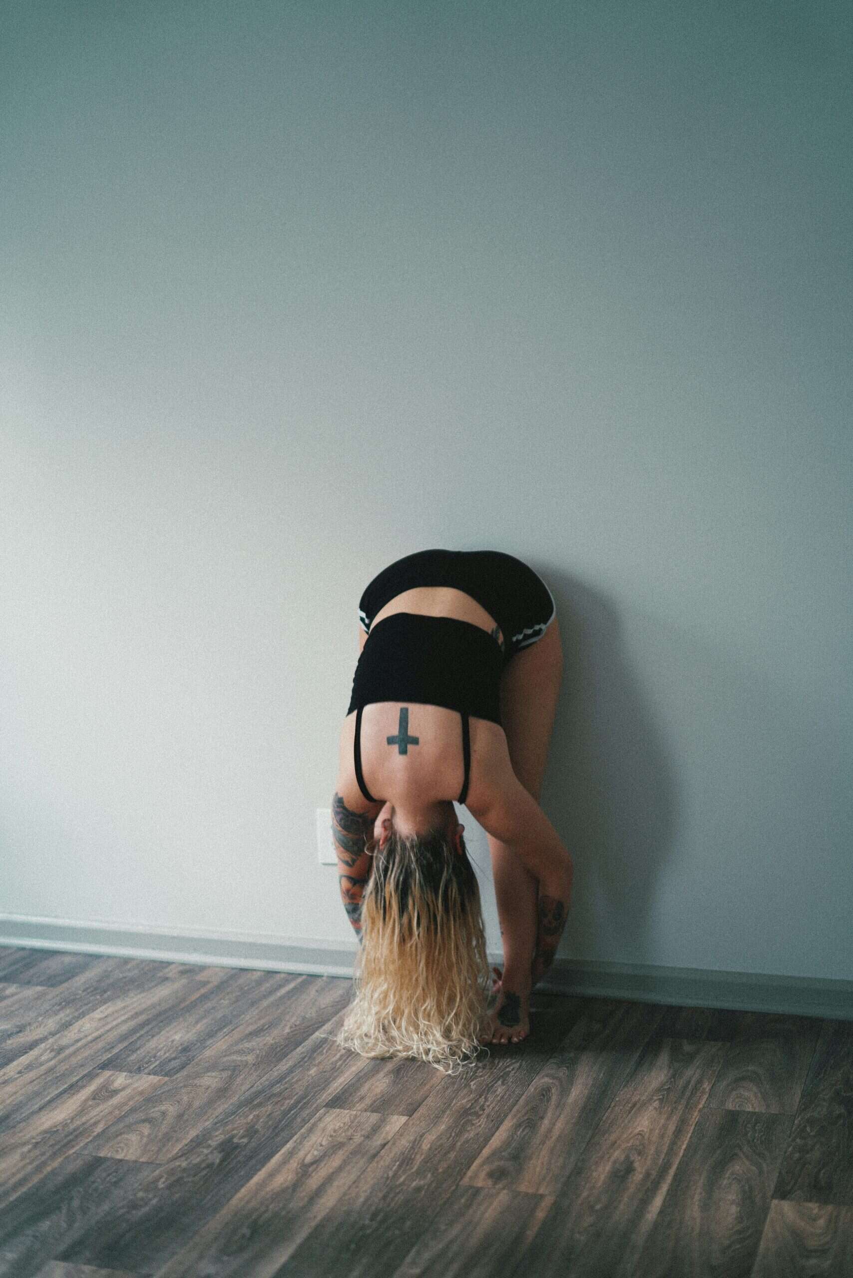 ron-mcclenny-tCKkGNgjUjc-unsplash Woman performing a wall roll down stretch, bending forward with her back to a plain wall in a minimal indoor space.