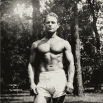 Vintage black-and-white photo of a muscular man standing shirtless outdoors wearing white shorts, embodying the early 20th-century fitness pioneer Joseph Pilates.