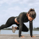 Woman performing a mountain climber exercise on a wooden dock by a lake, wearing black athletic wear and white sneakers, with a focused expression during an outdoor summer workout.