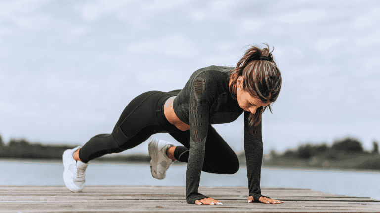 Woman performing a mountain climber exercise on a wooden dock by a lake, wearing black athletic wear and white sneakers, with a focused expression during an outdoor summer workout.