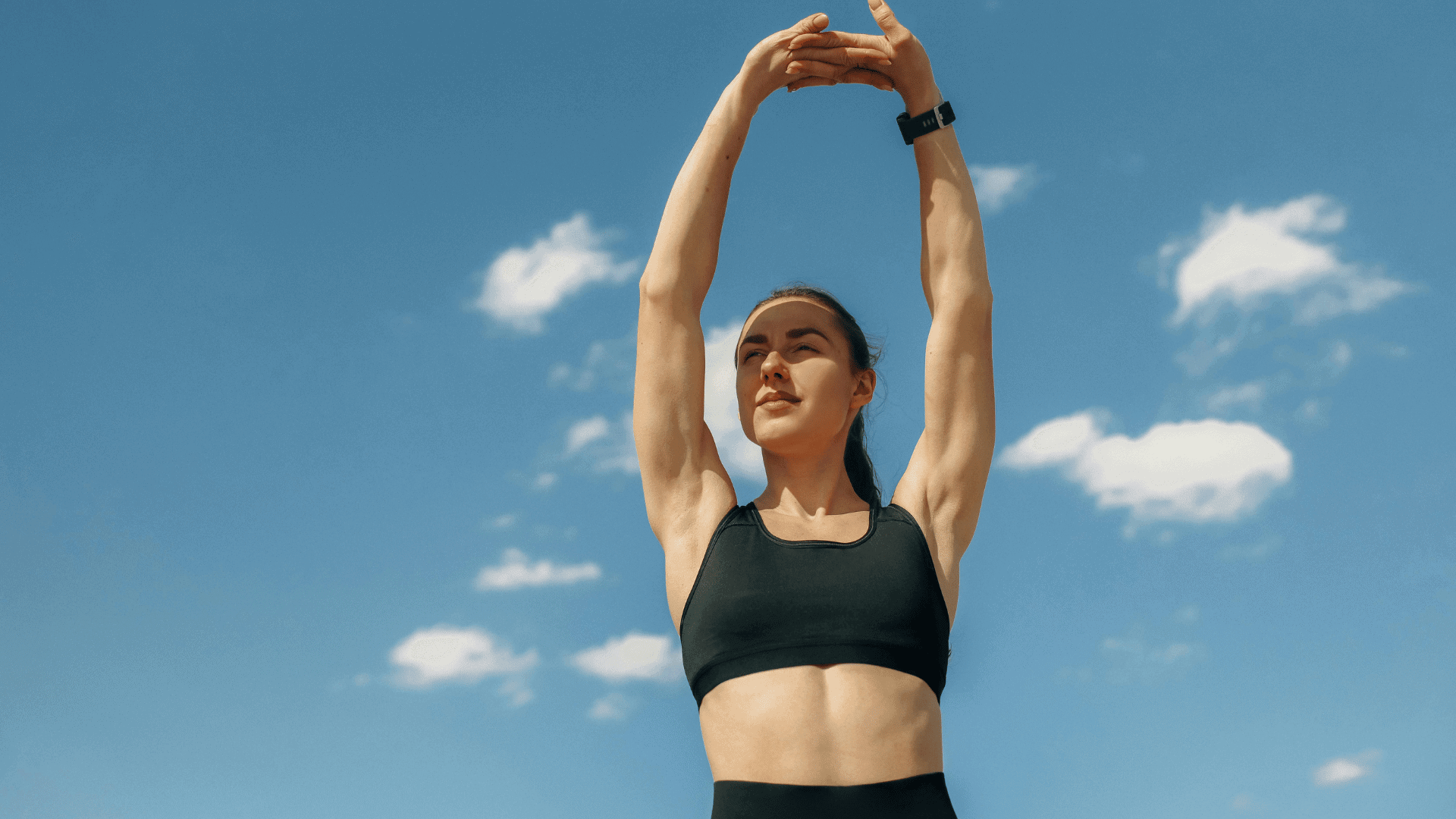 Confident athletic woman stretching with arms overhead against a clear blue sky, wearing a black sports bra and fitness watch, enjoying an energizing outdoor summer workout.