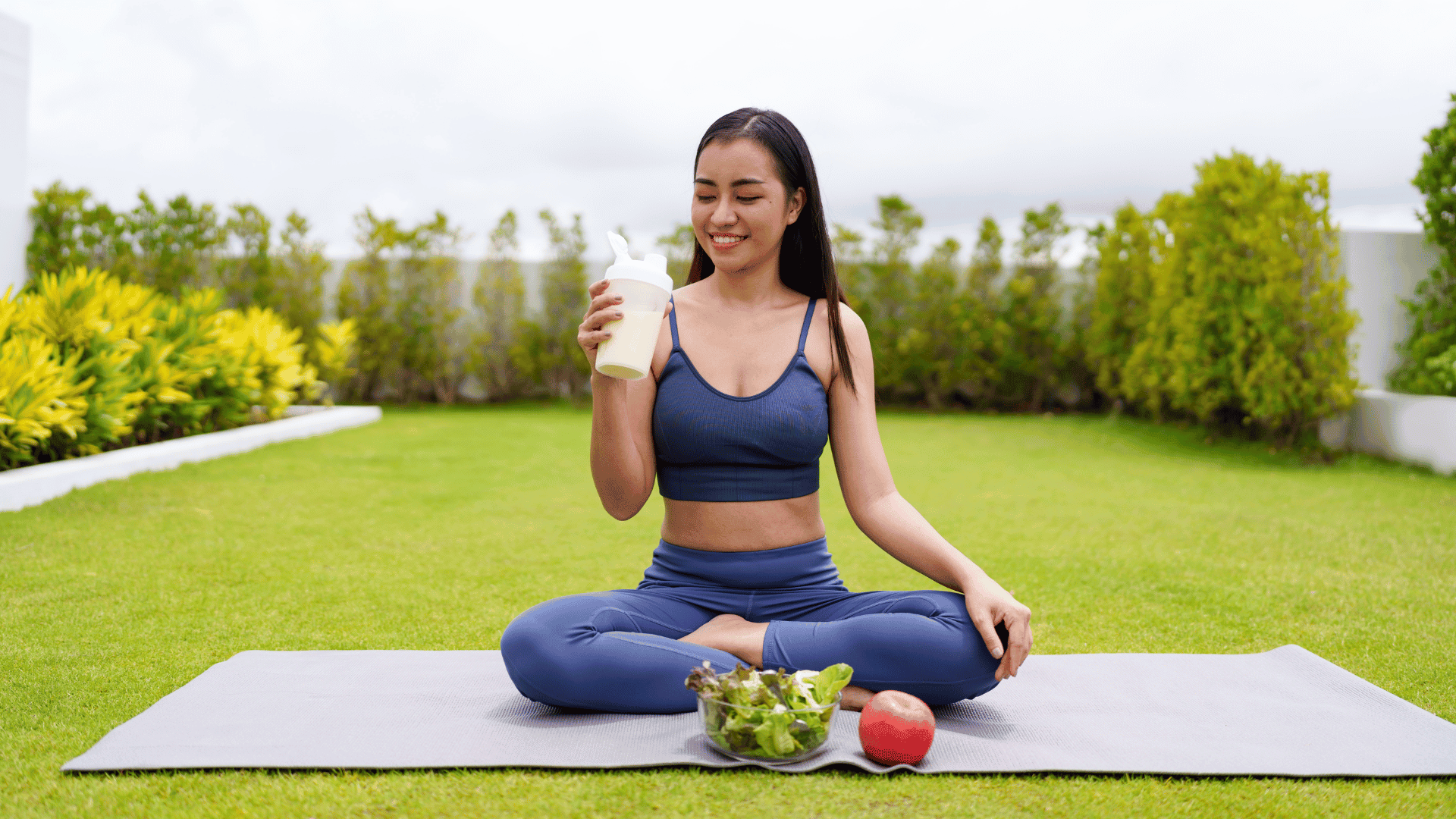 AN Overlay Template - 2025-08-06T145044.736 Woman sitting on a yoga mat outdoors, enjoying a protein shake with a bowl of salad and an apple — representing healthy post-workout nutrition to support muscle recovery and gains. Ask ChatGPT
