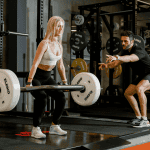 Woman lifting a barbell under the guidance of a personal trainer at the gym, illustrating a structured workout plan to tone and strengthen the body.