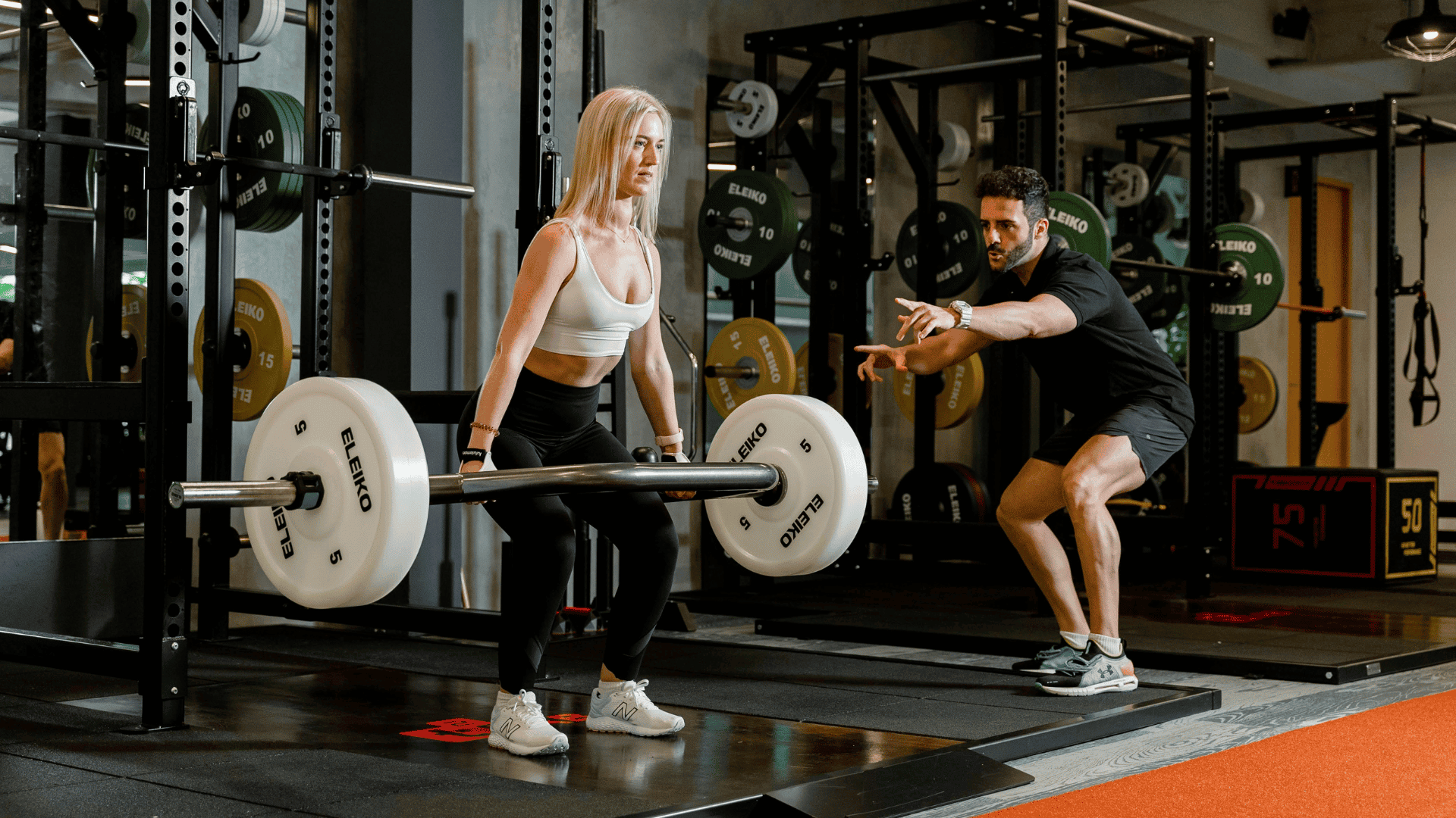 Woman lifting a barbell under the guidance of a personal trainer at the gym, illustrating a structured workout plan to tone and strengthen the body.