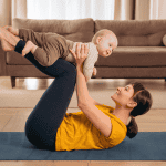 Mother lying on a yoga mat at home, lifting and smiling at her baby during a gentle postpartum workout.