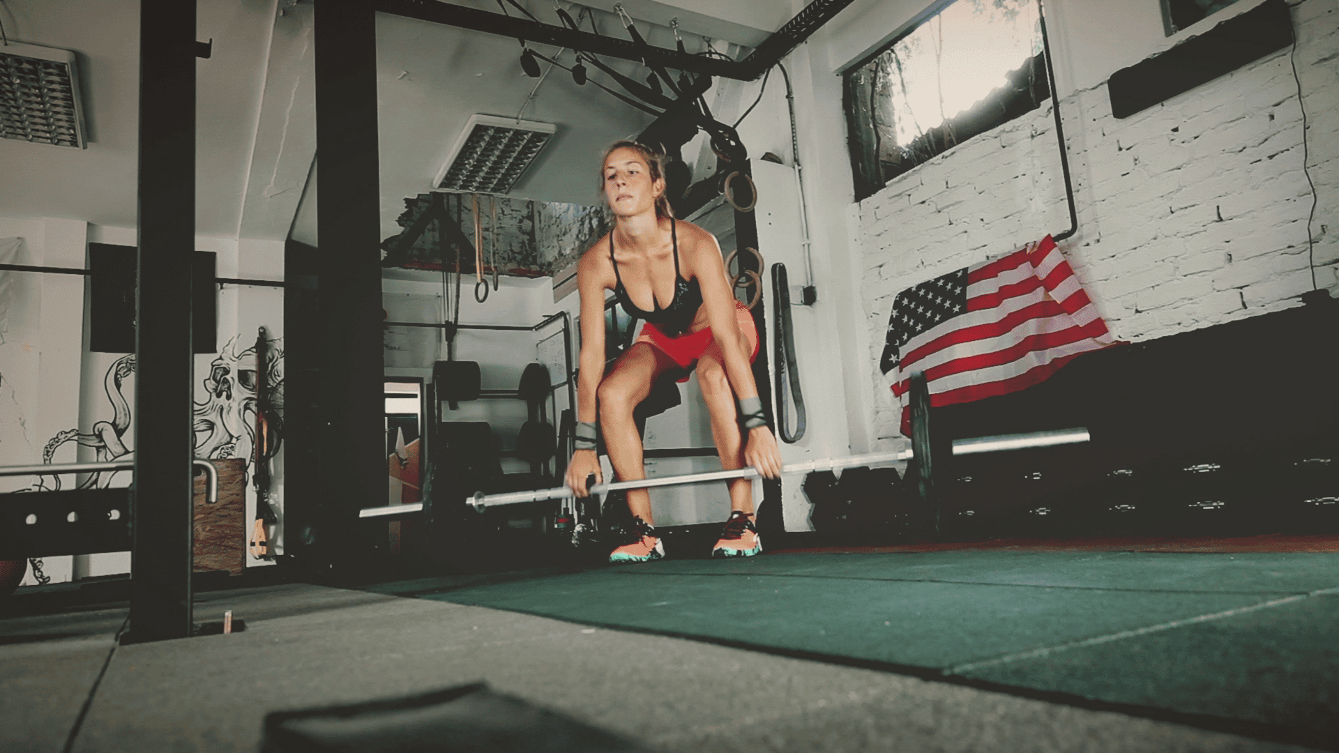 Woman preparing to lift a barbell in a gym with an American flag in the background.