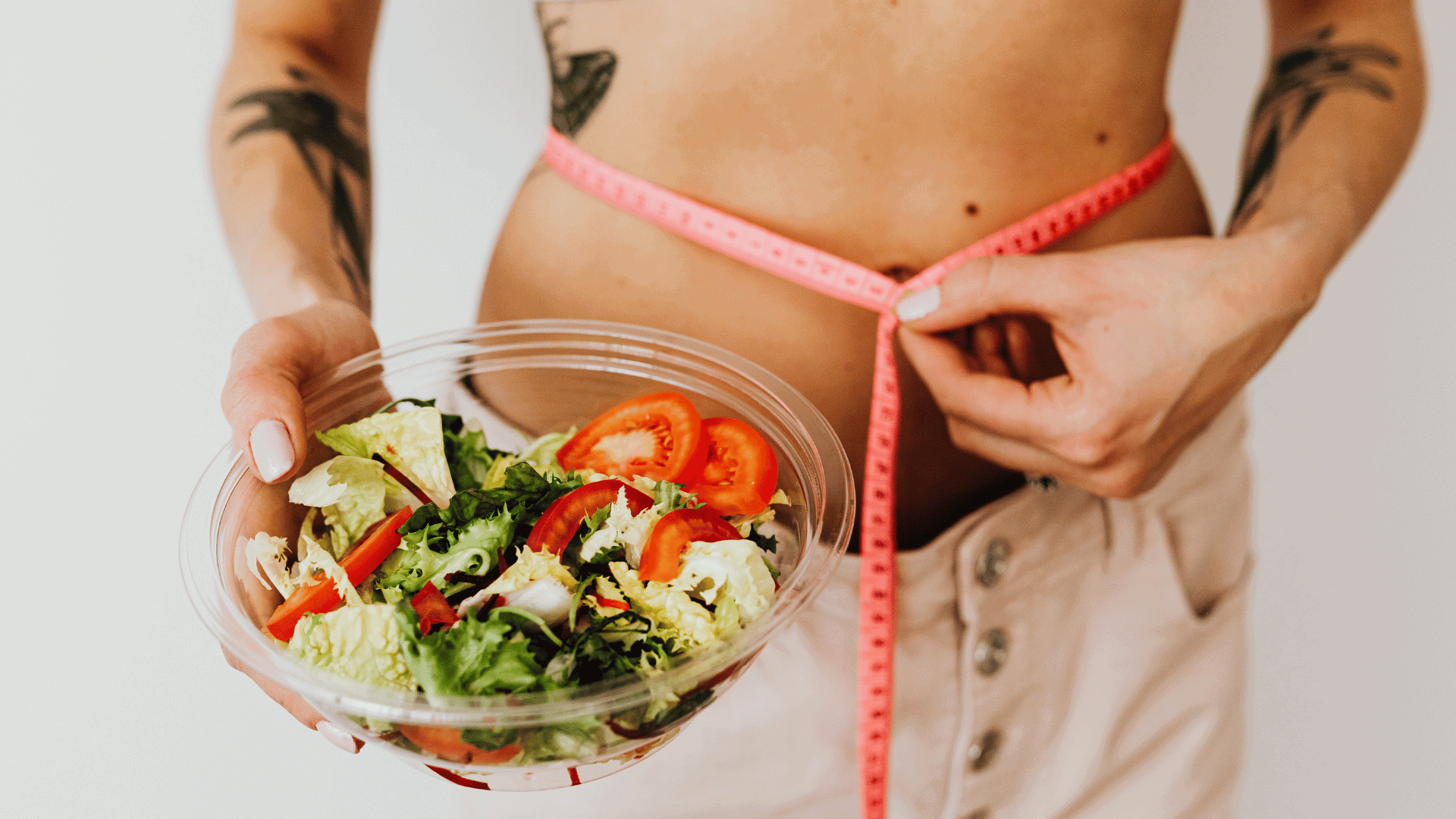 Woman holding a bowl of fresh salad while measuring her waist with a tape measure, emphasizing healthy eating for fat loss.