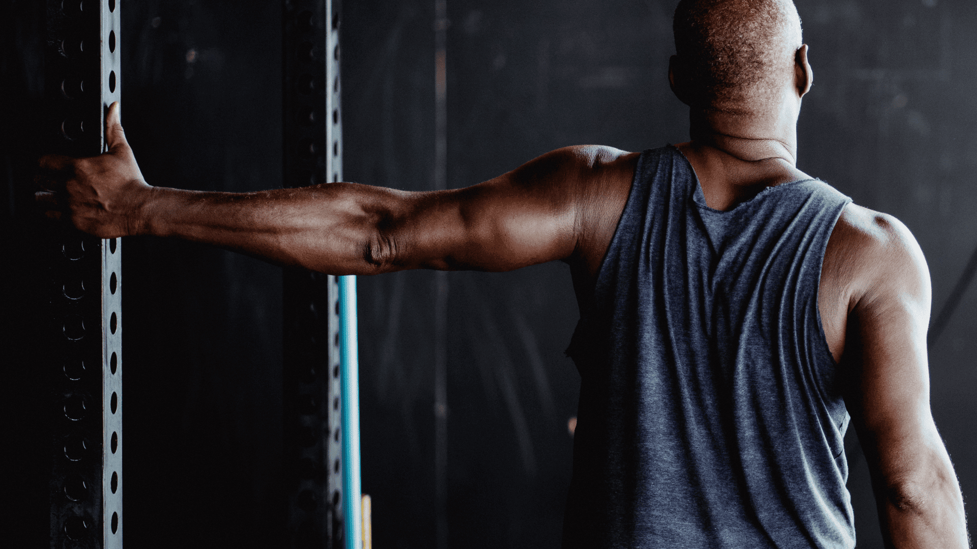 Man in a sleeveless shirt stretching his shoulder by holding onto a gym rack, viewed from behind, highlighting the importance of posture and core engagement during workouts.