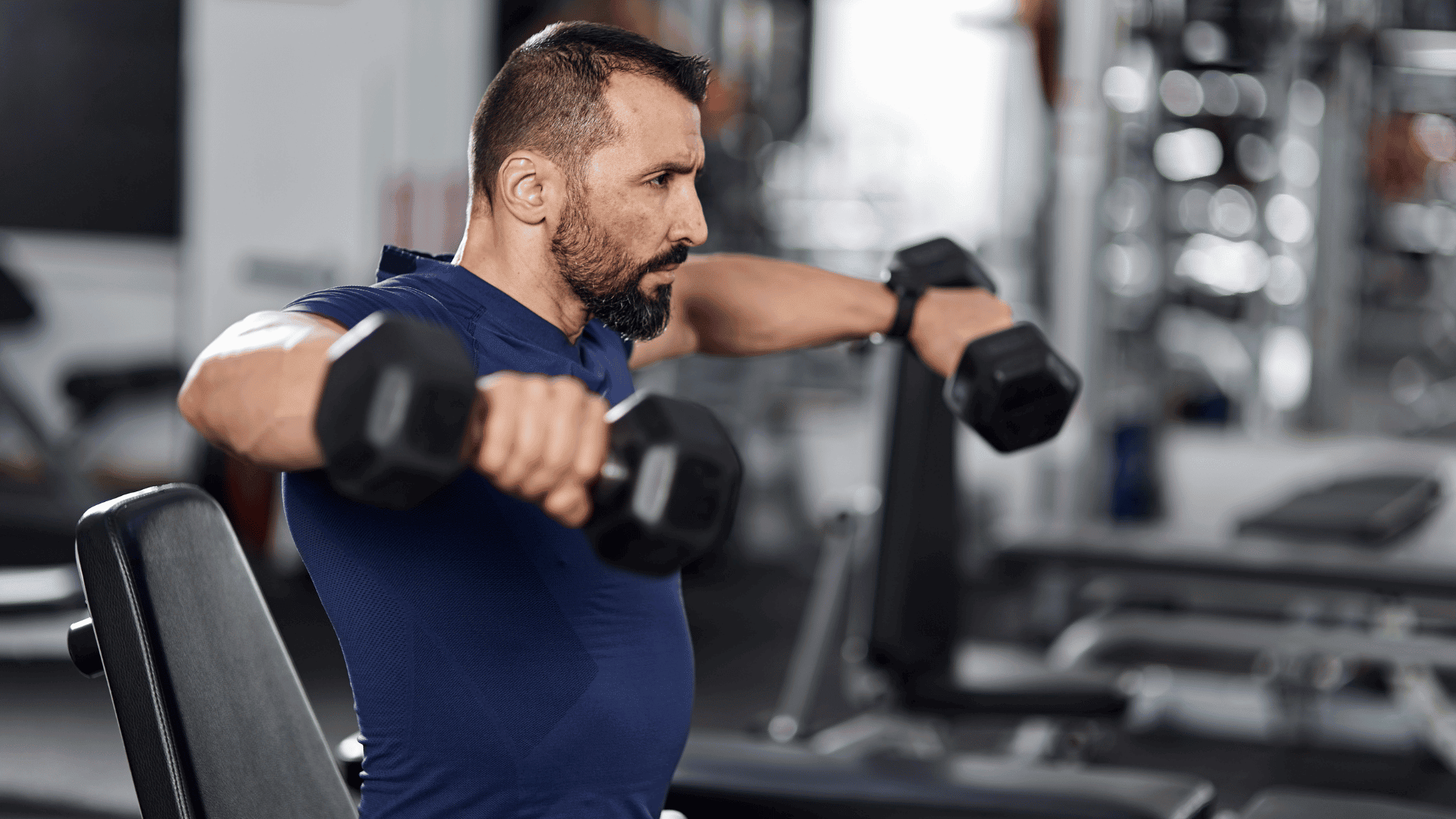 ocused man in a navy blue athletic shirt performing seated dumbbell lateral raises in a gym, emphasizing steady volume and consistent shoulder training.