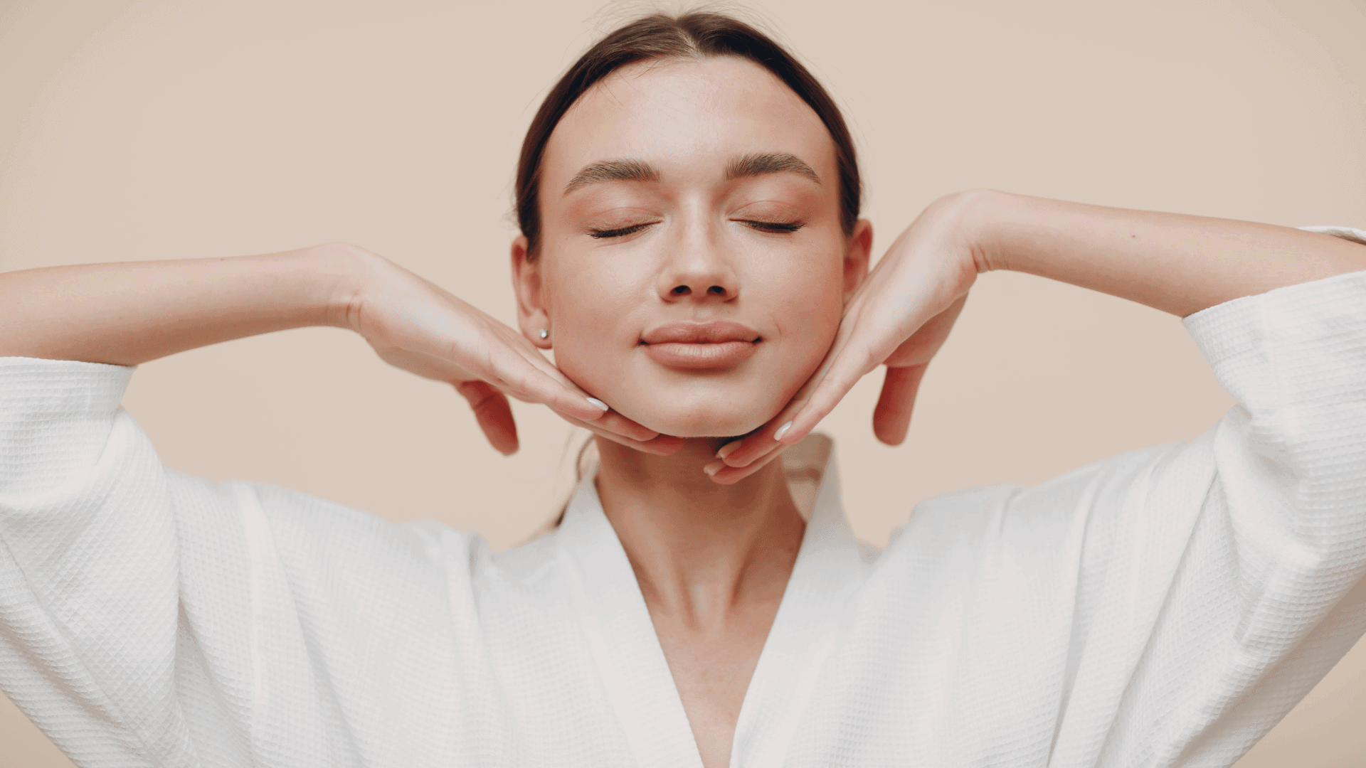 Young woman practicing face yoga with her hands gently placed under her chin, eyes closed, wearing a white robe against a neutral background, promoting facial relaxation and toning.