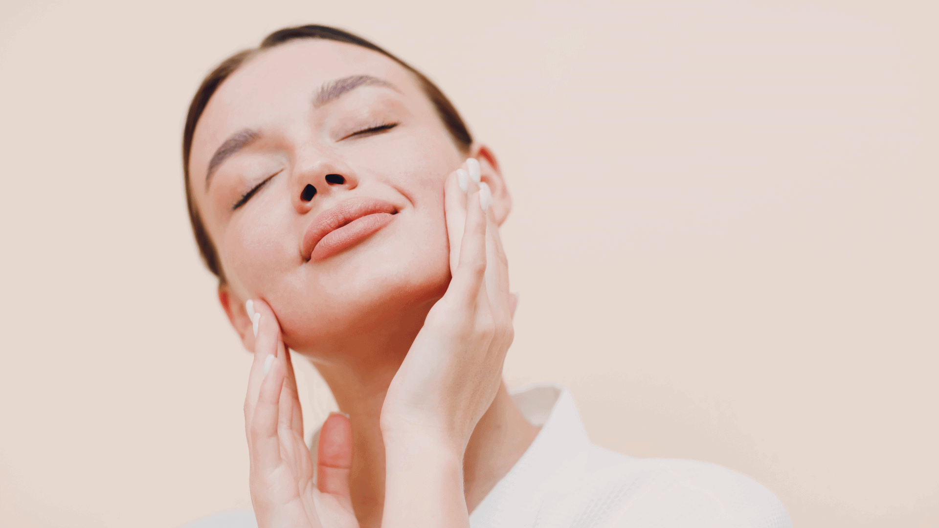 Close-up of a woman practicing face yoga with a relaxed expression, gently touching her cheeks with both hands, eyes closed, highlighting skincare and self-care benefits.