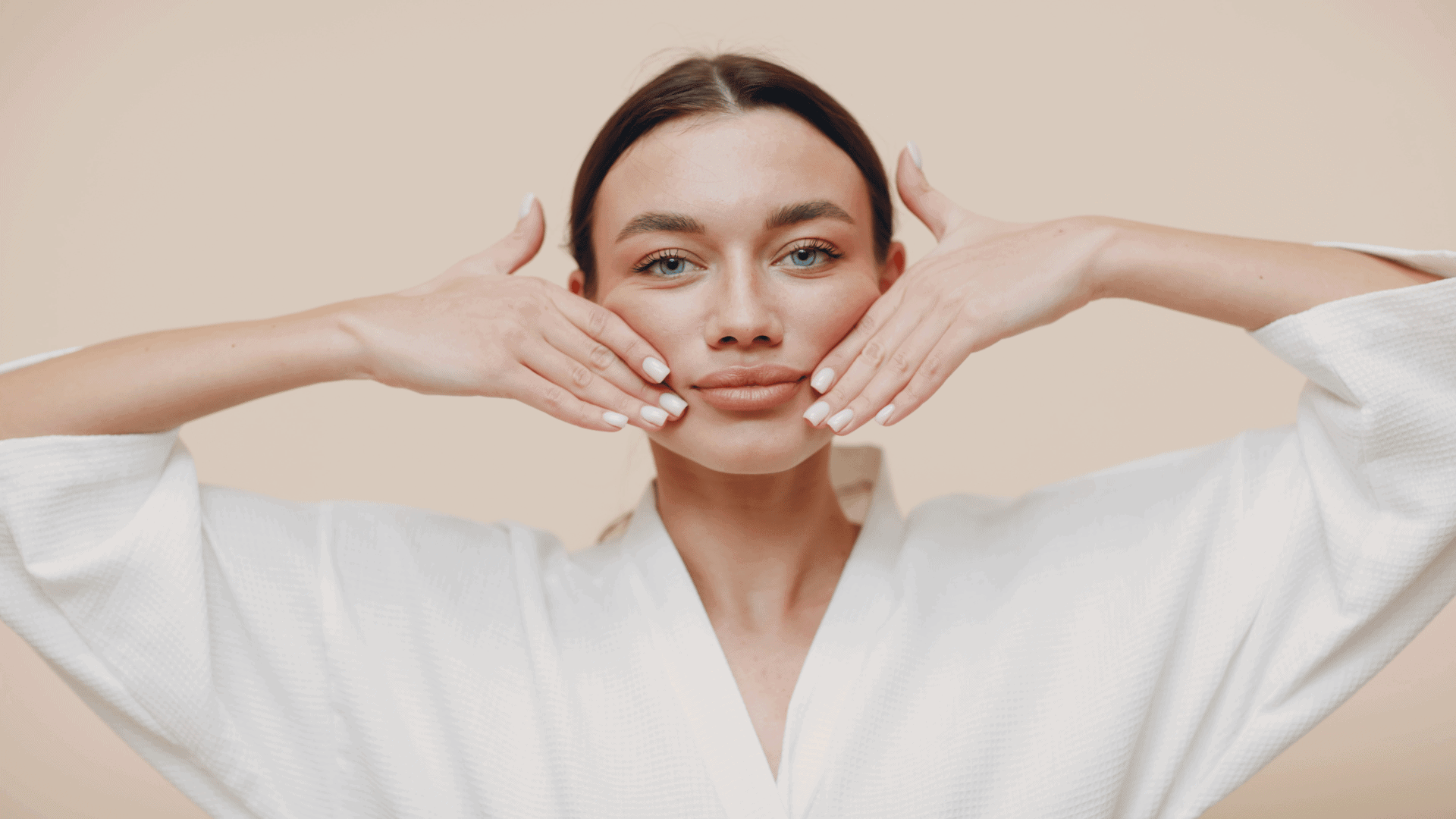 Young woman in a white robe performing the 'Happy Cheeks Sculpting' face yoga exercise, pressing her fingertips gently into the cheeks while smiling to activate and lift the cheek muscles.