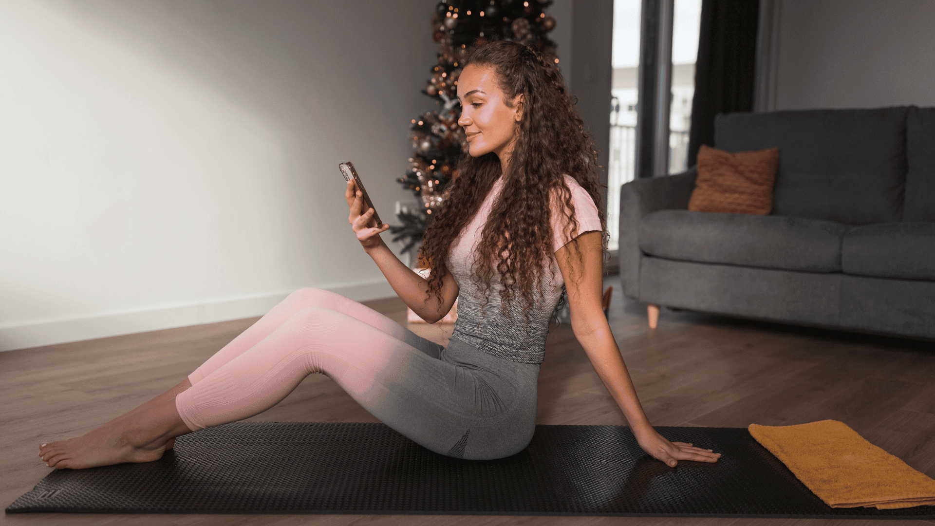 Young woman sitting on a yoga mat in a cozy home with a Christmas tree in the background, casually looking at her phone post-workout, appearing distracted.